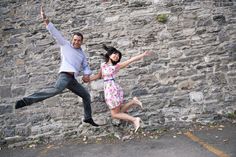 a man and woman jumping in the air with their arms spread wide, against a stone wall