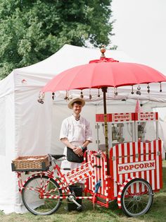 a man standing next to a red and white food cart with an umbrella over it