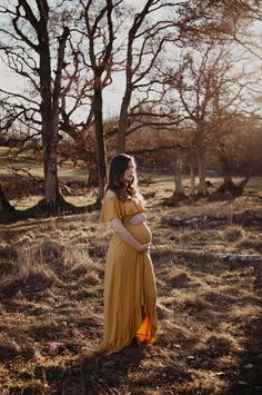 a pregnant woman wearing a yellow dress standing in an open field with bare trees behind her