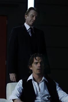 two men in suits and ties sitting on a chair with one man standing behind them