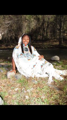 a woman sitting on the ground next to a river with rocks and grass around her