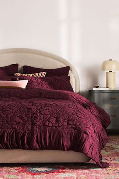 a bed with purple comforter and pillows in a room next to a lamp on a dresser