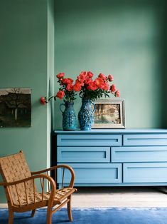 two vases with red flowers sit on a blue dresser next to a wooden chair