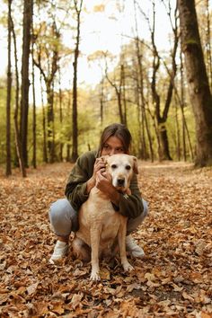 a woman kneeling down holding a dog in the woods with leaves on the ground around her