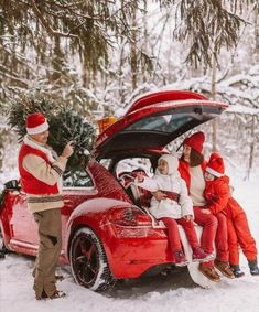 a group of people sitting on top of a red car in the snow next to a christmas tree