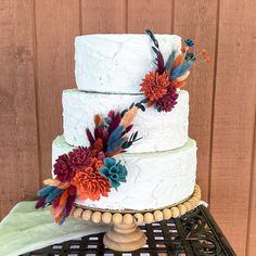 a three tiered cake with flowers on top sits on a table in front of a wooden wall