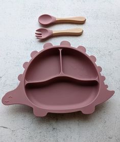 a pink dinosaur shaped divided plate with spoons and fork next to it on a white surface