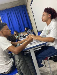 two young men sitting at a table in front of a laptop computer and talking to each other