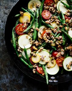 a skillet filled with green beans, tomatoes and other veggies on top of a table