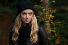 a woman with long blonde hair wearing a black hat and jacket standing in front of trees