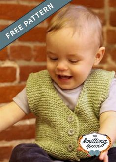 a baby wearing a green knitted vest sitting on top of a wooden floor next to a brick wall