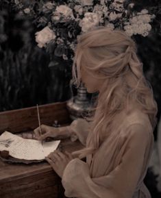 a woman sitting at a table writing on a piece of paper with flowers in the background