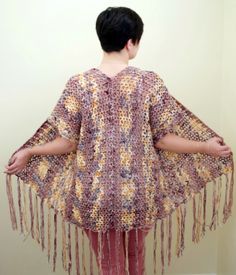 a woman standing in front of a wall wearing a crocheted shawl with tassels