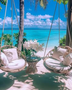 two chairs and a table on a beach with palm trees in the foreground, blue water in the background