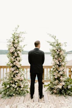 a man in a black suit standing on a wooden deck next to flowers and greenery