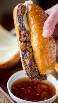 a hand holding a sandwich over a bowl of sauce on a tray with bread in the background