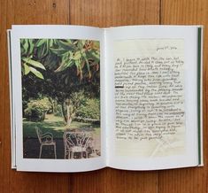 an open book on a wooden table with chairs and trees in the backgroud