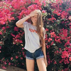 a girl standing in front of pink flowers with her hands on her head and smiling