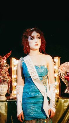 a woman with red hair wearing a blue dress and holding a white sash in front of a mirror