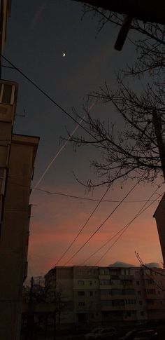 the sun is setting behind some buildings and power lines in the foreground, with an airplane streaking across the sky