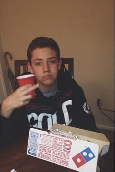 a young man sitting at a table with a pizza box and cup in front of him