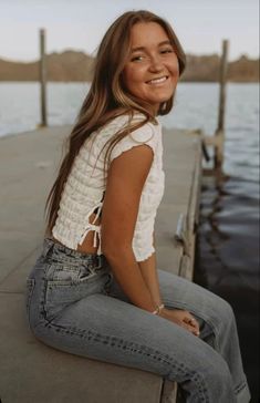 a woman is sitting on a dock smiling at the camera while wearing jeans and a white top