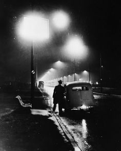 a black and white photo of a man standing in the rain next to an old car