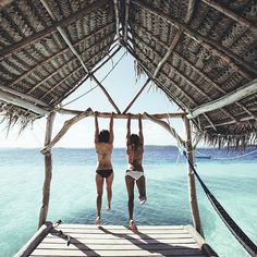 two women jumping off a dock into the ocean from an overhanged hut with thatched roof