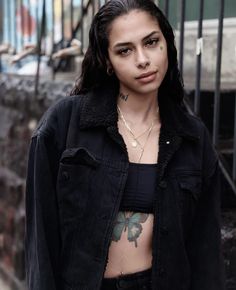 a woman with tattoos on her chest standing in front of a fenced area and looking at the camera