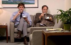 two men sitting on couches eating food in an office setting, one is holding papers and the other is reading