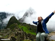 a woman sitting on top of a mountain with her arms in the air and clouds behind her