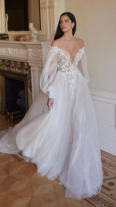 a woman standing in front of a fireplace wearing a wedding dress with long sleeves and an open back