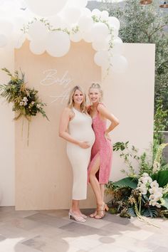 two women standing next to each other in front of a wall with balloons and greenery