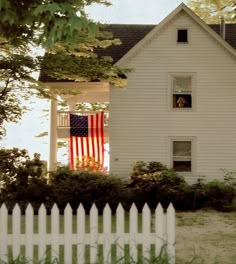 a white house with an american flag hanging from the front window and a picket fence surrounding it