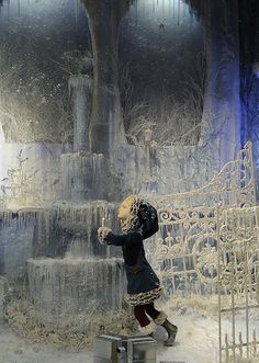 a woman in winter clothing standing next to an ice - covered fountain with snow on the ground