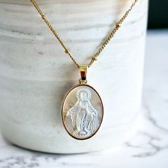 Oval shaped gold Virgin Mary pendant necklace featuring mother of pearl.  Material: Gold plated with cubic zirconia crystals. Oval Shape. Pendant size: 1" x  .5" Chain Length 17" *Satin Gift Bag Included 1. Personal Expression of Faith: Wearing this necklace serves as a personal and outward declaration of one's faith. It can act as a daily reminder of one's spiritual beliefs, values, and connection to God. For the individual wearing it, this Christian necklace can also serve as a touchpoint for White Oval Gold Plated Necklace, White Oval Gold-plated Necklace, White Gold-plated Necklaces For Mother's Day, Elegant Miraculous Medal Necklace, White Gold Plated Necklaces For Mother's Day, White Medallion Necklace For Anniversary, Silver Oval Pendant Charm Necklace In Gold Plated, Gold Medallion Mother Of Pearl Jewelry, Gold Medallion Jewelry With Mother Of Pearl