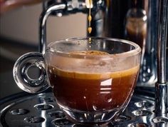 coffee being poured into a glass cup on top of a metal tray with water in it