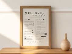 a framed welcome sign sitting on top of a wooden table next to a vase and wall