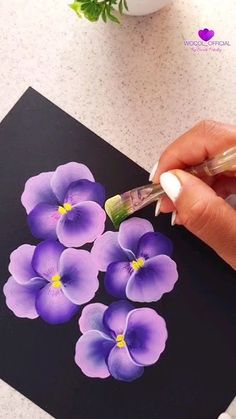 a woman is painting purple flowers on a black piece of paper with a brush and acrylic paint