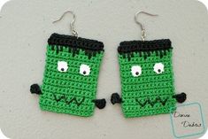 two green crocheted monster earrings with black eyes and ears, one is made to look like a mug