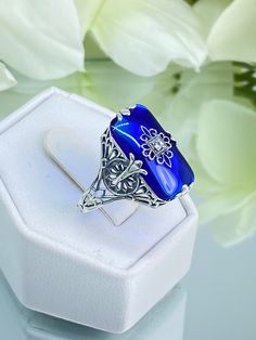 Sapphire Blue Glass Sterling Silver Ring Grace Design#D233 This is a lovely antique Edwardian era inspired filigree ring. This gorgeous ring is created in stunning sapphire blue glass. The glass is embellished with a delicate floral filigree. The top of the ring holds a center set 1mm round cut gemstone (Choose from white Cubic Zirconia, lab created white Moissanite, or Natural/Genuine White Diamond). The blue glass is 18mm x 19mm. The ring sits 18mm north-south and 10mm east-west on the finger. The inside of the band is marked 925 for sterling silver. The band's intricate open work filigree has hints of floral influence and geometric design running its entire length. The curves and swirls accent the beautiful smooth stone. The center filigree highlights the sparkling gemstone. This ring r Blue Intaglio Ring As Gift, Vintage Blue Filigree Ring For Anniversary, Vintage Blue Filigree Rings, Vintage Blue Sapphire Ring With Intricate Design, Sapphire Victorian Rings For Gift, Vintage Blue Filigree Ring For Wedding, Victorian Sapphire Rings As Gift, Vintage Blue Filigree Ring As Gift, Ornate Filigree Ring With Stone Setting