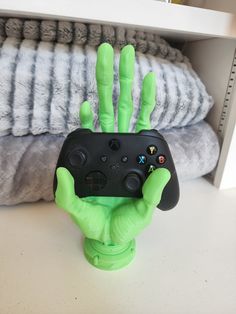 a green hand holding a black controller on top of a white table next to pillows