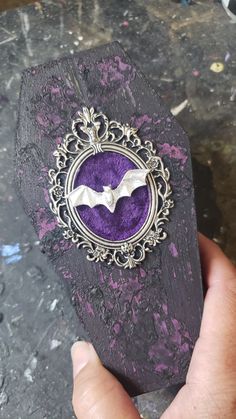 a hand holding a purple bat brooch on top of a piece of wood with filigreet around it