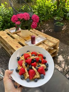 a person holding up a bowl of cereal with berries on it and pink flowers in the background