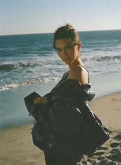 a woman standing on top of a beach next to the ocean holding a black bag