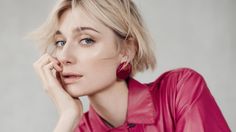 a woman with short blonde hair wearing a pink shirt and earrings, posing in front of a gray background