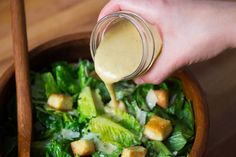 a person pouring dressing into a salad in a bowl with lettuce and croutons