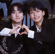 two young men sitting next to each other making a heart shape with their hands in front of them