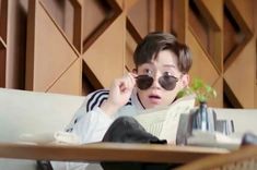 a boy wearing sunglasses reading a book while sitting on a couch with a plant in front of him