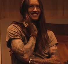 a young man with long hair and tattoos on his arms is smiling at the camera